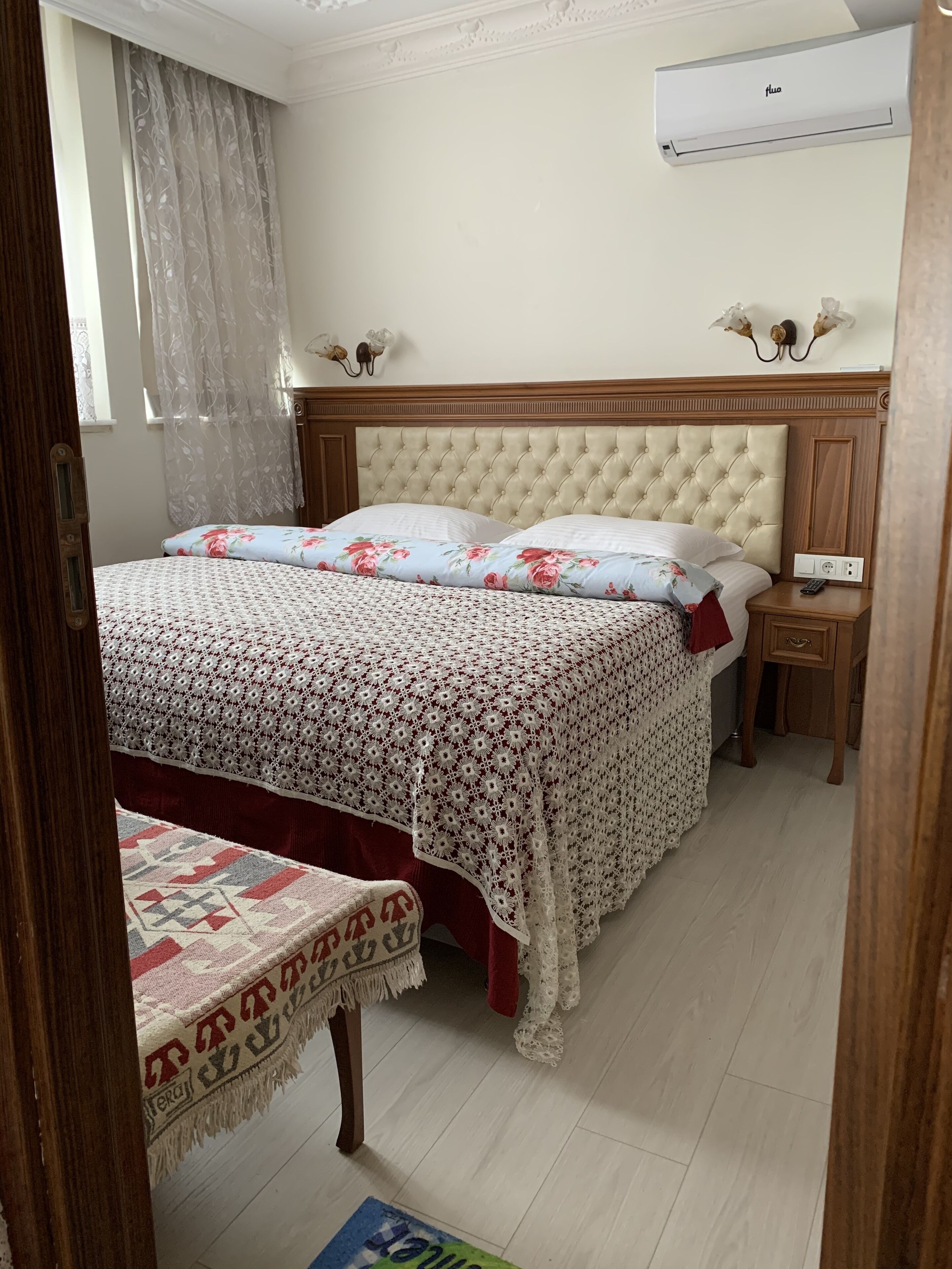 A bedroom with a large bed, a bedside table, a nightstand, an air conditioning unit, and a window with curtains.