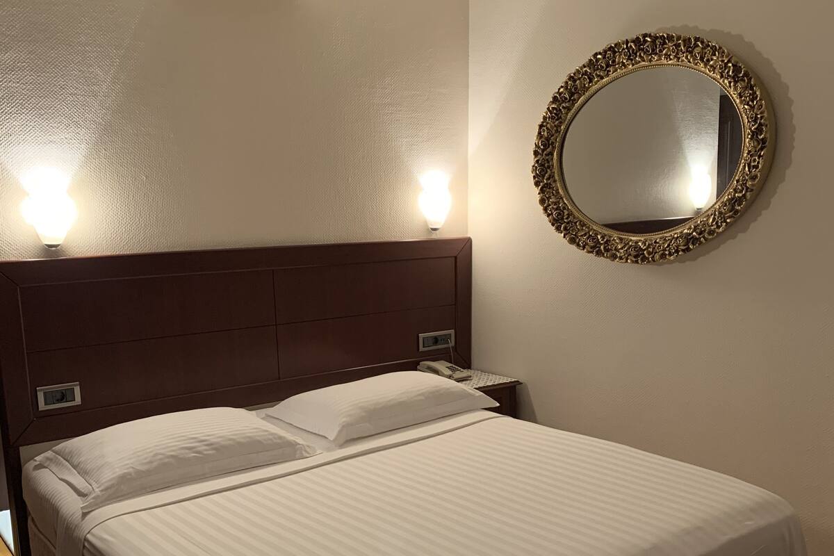 A neatly made bed with a headboard, a wall-mounted mirror, and an air conditioning unit above it.