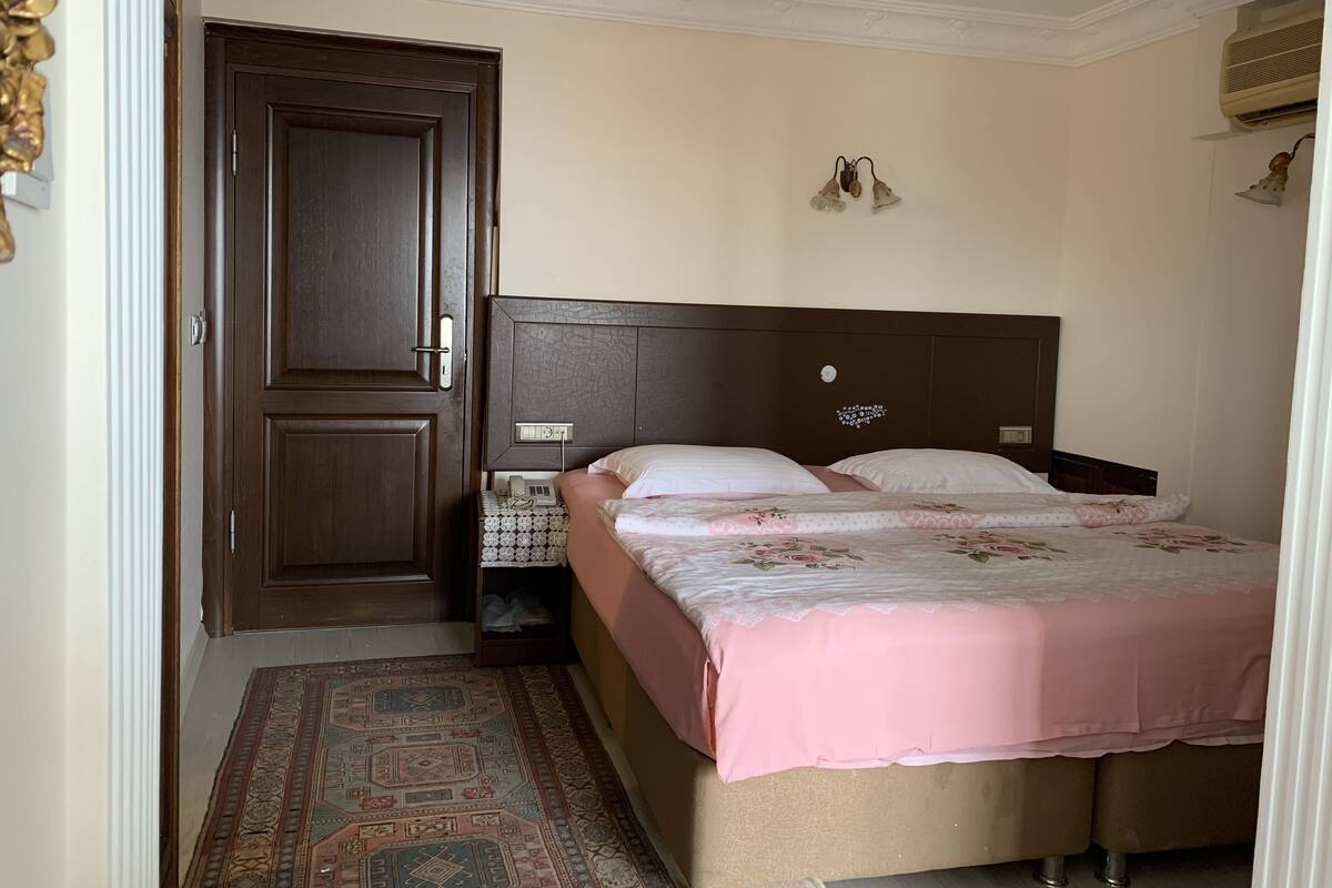 A bedroom with a double bed, a wooden door, a bedside table, and a patterned rug.