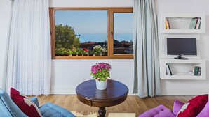 Een woonkamer met uitzicht op zee, een ronde houten tafel met een bloevas, een boekenkast en een televisie.