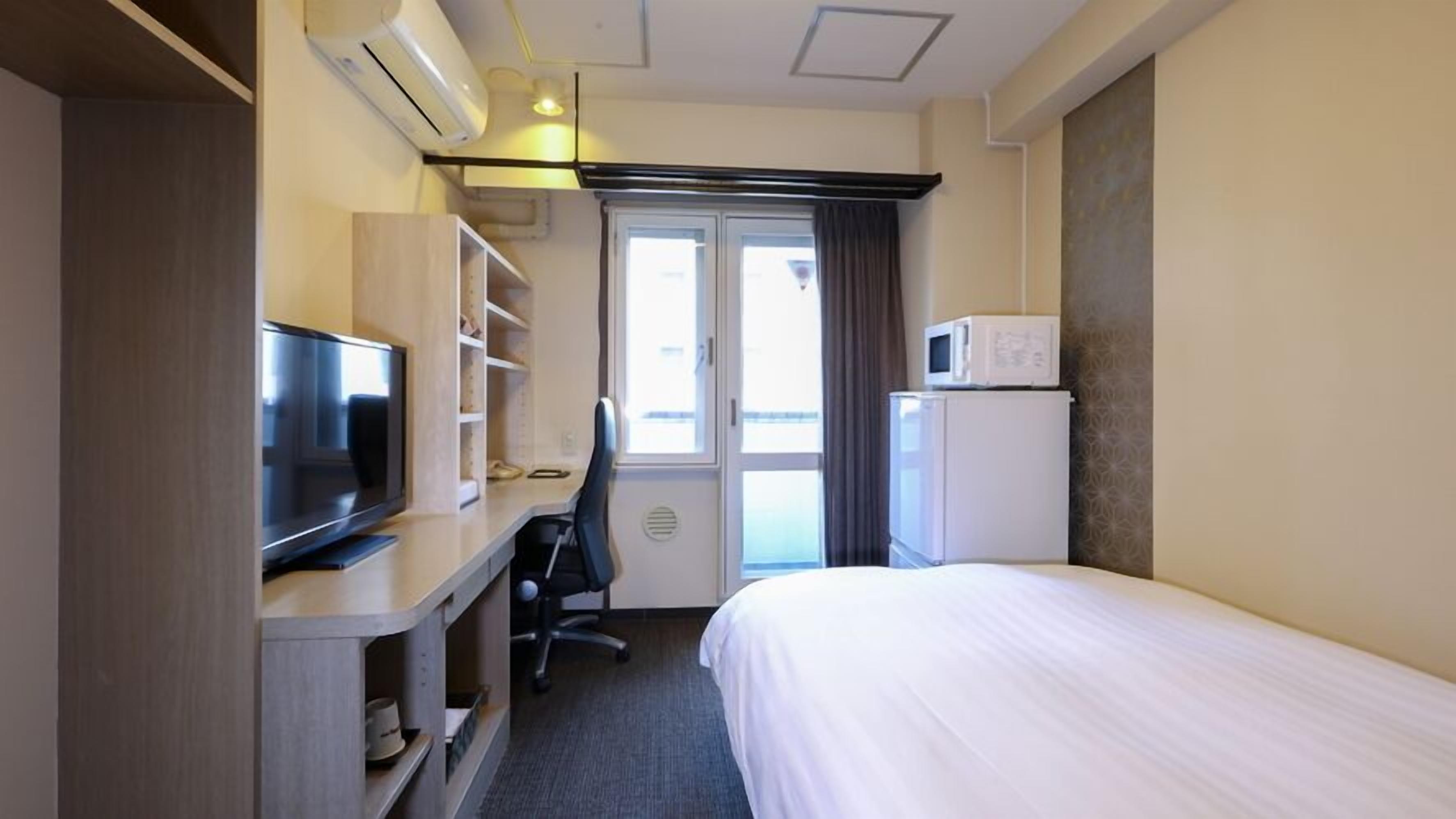 A hotel room with a bed, a desk with a television, a microwave, and a window with curtains.