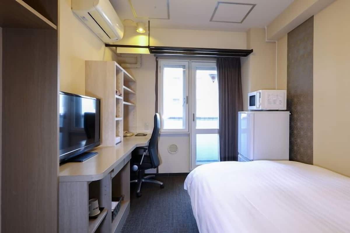 A hotel room with a bed, a desk with a television, a microwave, and a window with curtains.