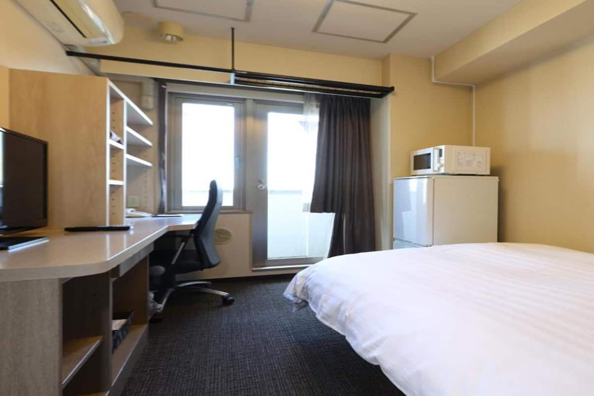 A hotel room with a bed, desk, chair, television, microwave, refrigerator, and a window with curtains.