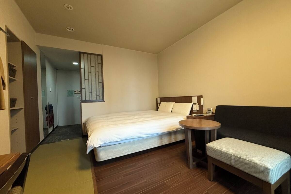 A compact hotel room with a bed, a small round table, a sofa, and a wooden floor.