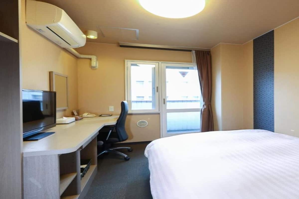 A hotel room with a bed, a desk with a chair, a television, and an air conditioning unit.