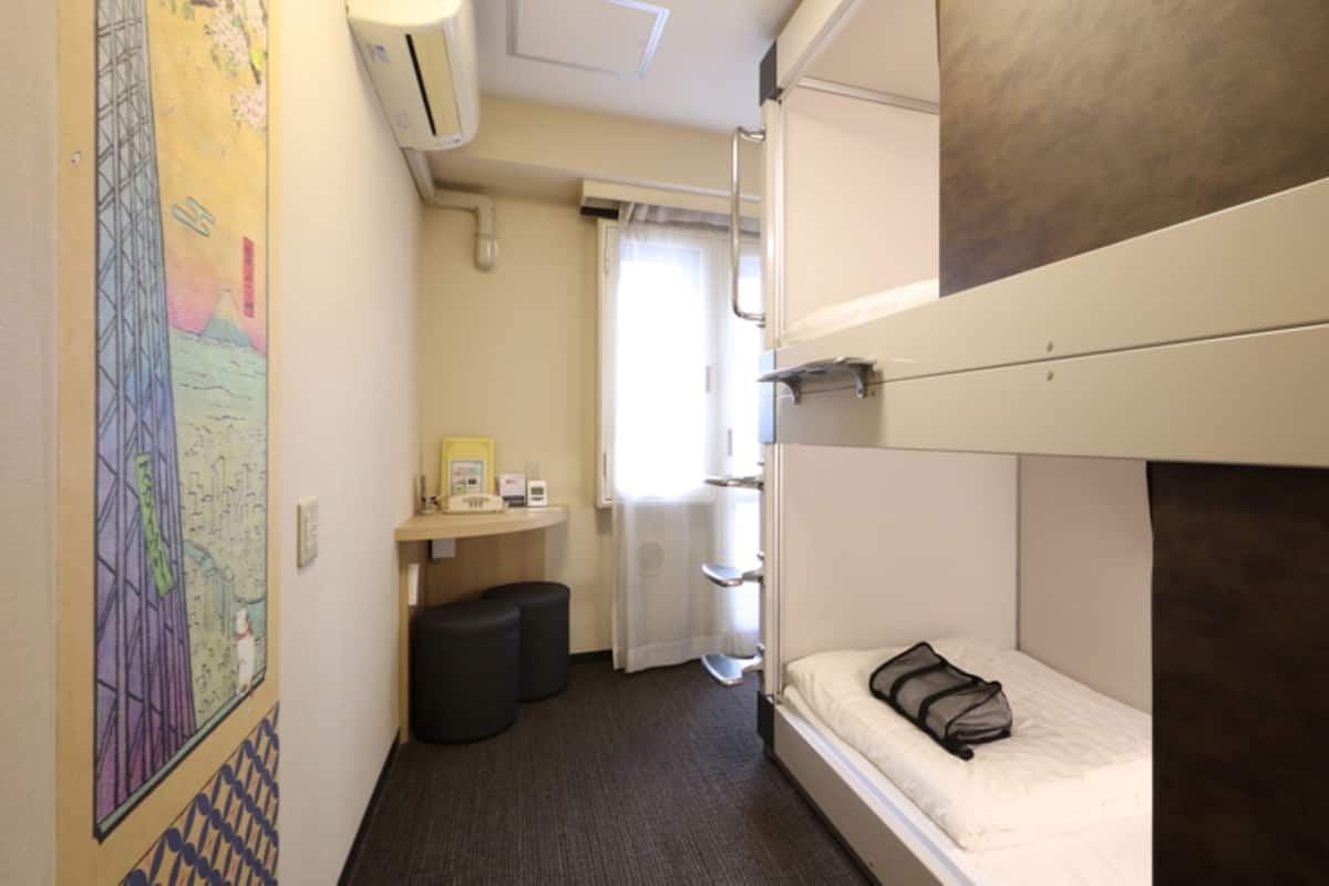 A compact hotel room with bunk beds, a small desk with a chair, a trash bin, and a window with curtains.