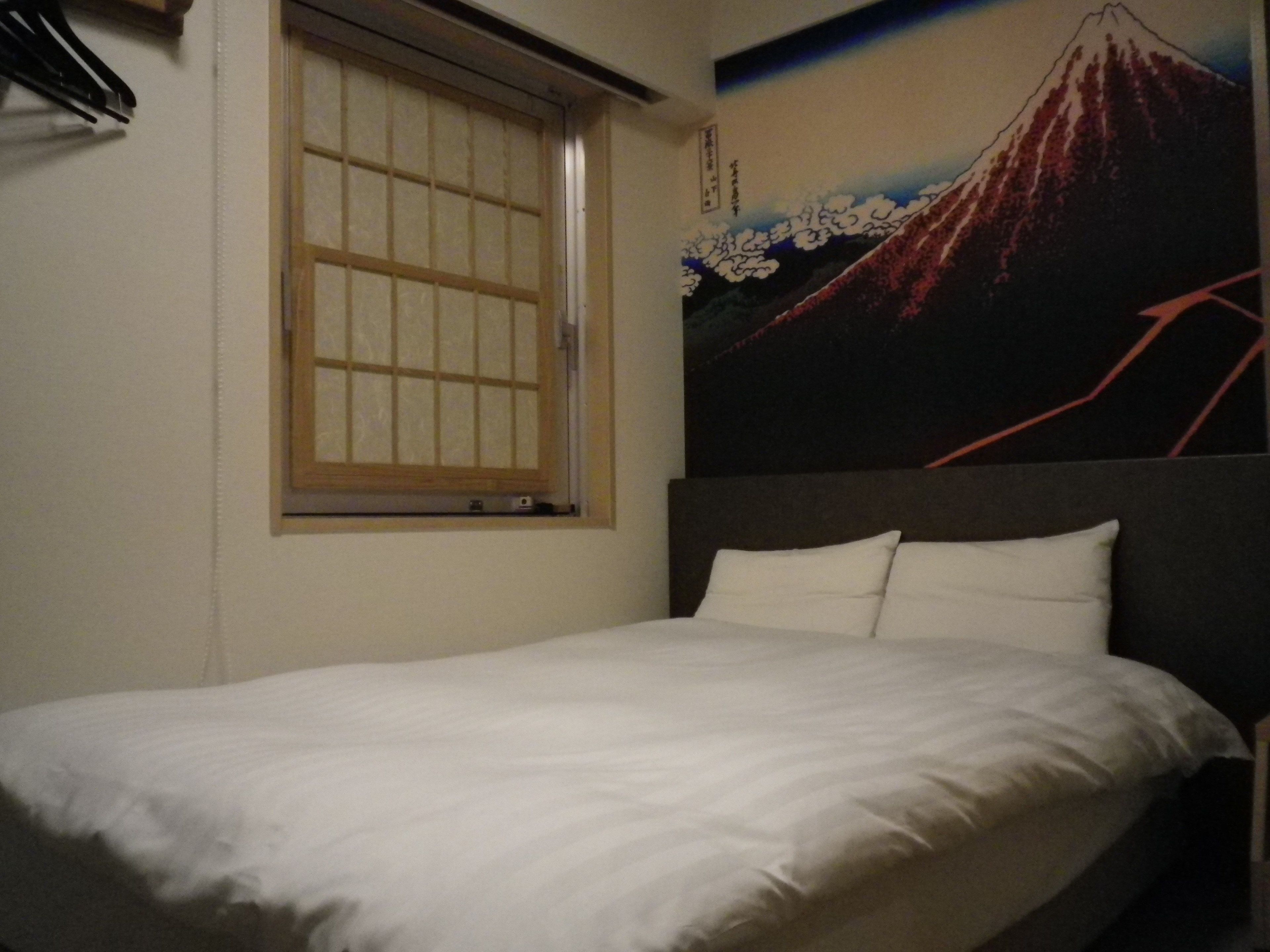 A bedroom with a bed, a window with a wooden frame, and a wall with a painting of a mountain.