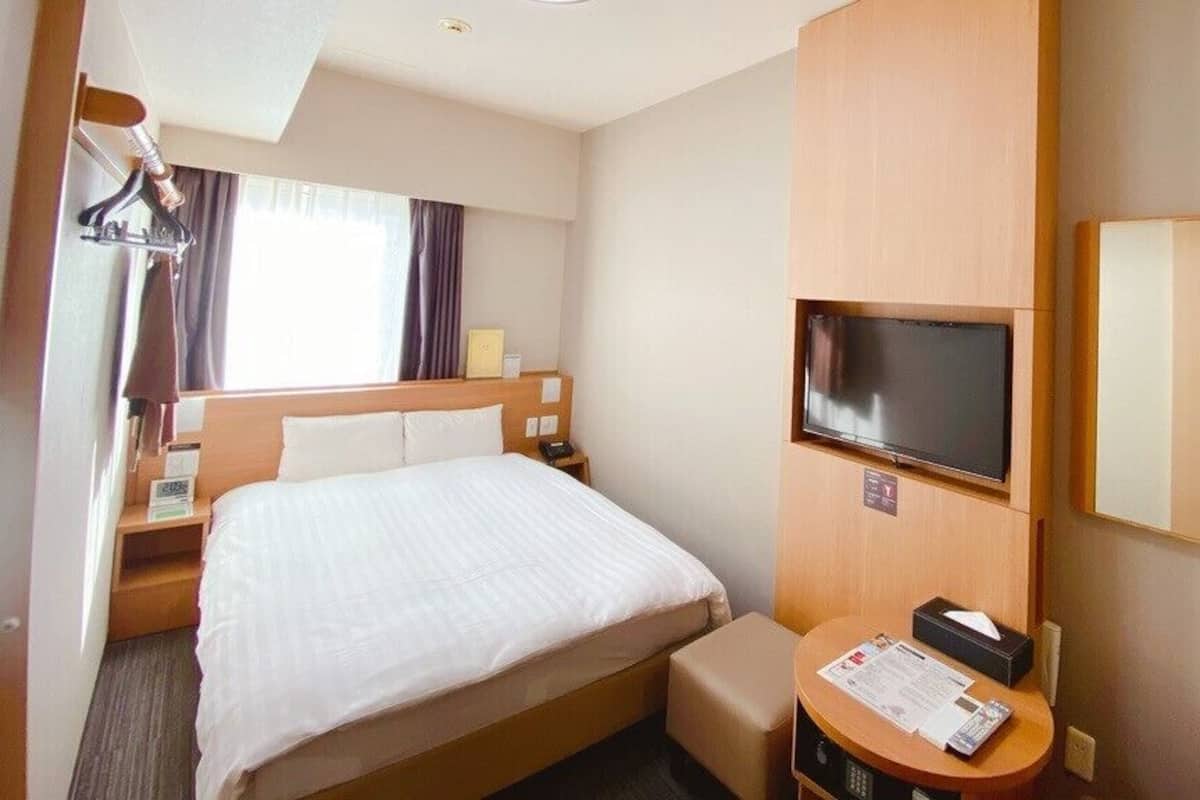 A hotel room with a bed, a TV, a desk with a chair, a mirror, and a window with curtains.