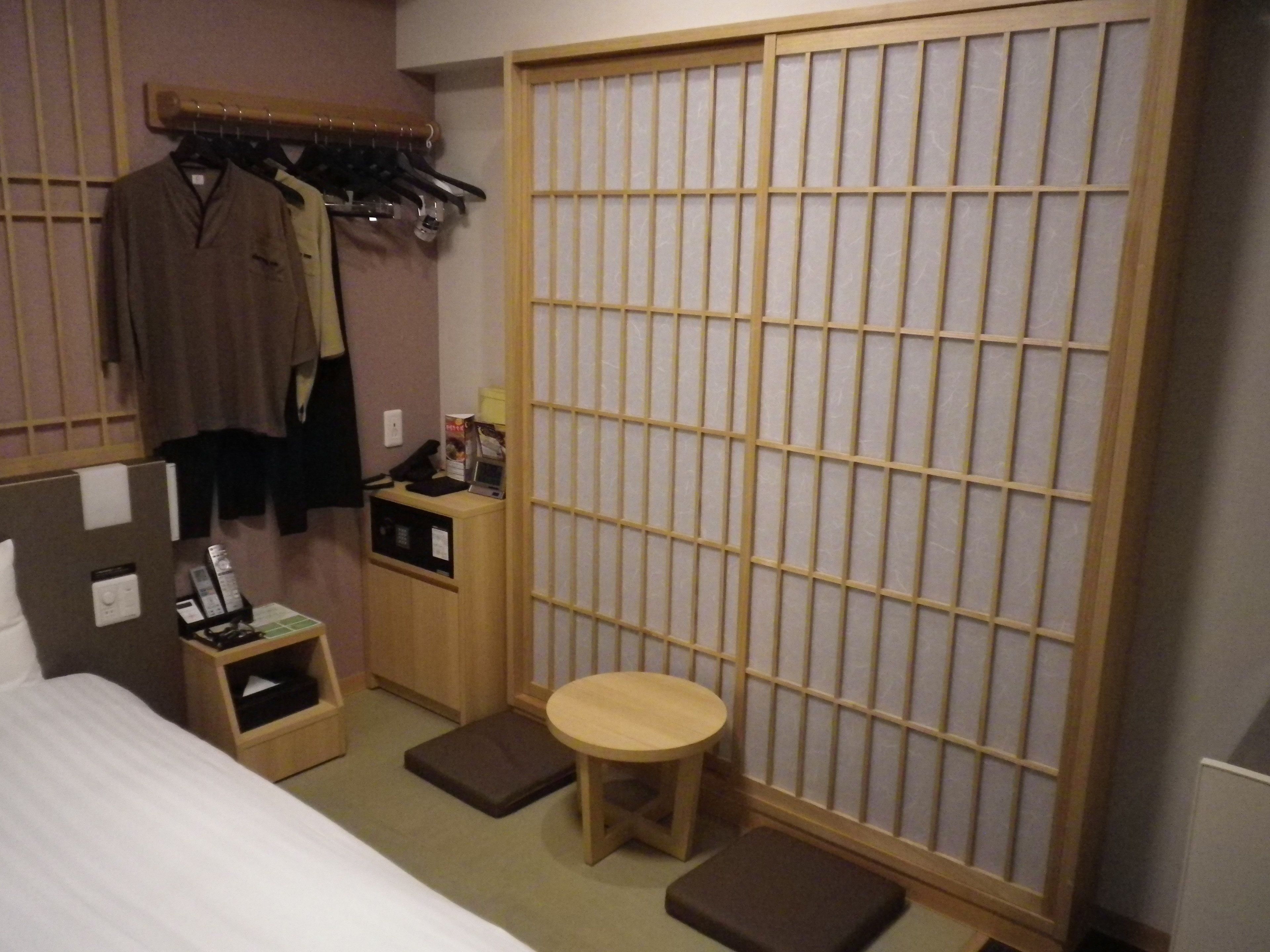 A hotel room with a bed, a small table, a stool, a wardrobe, and a sliding door.