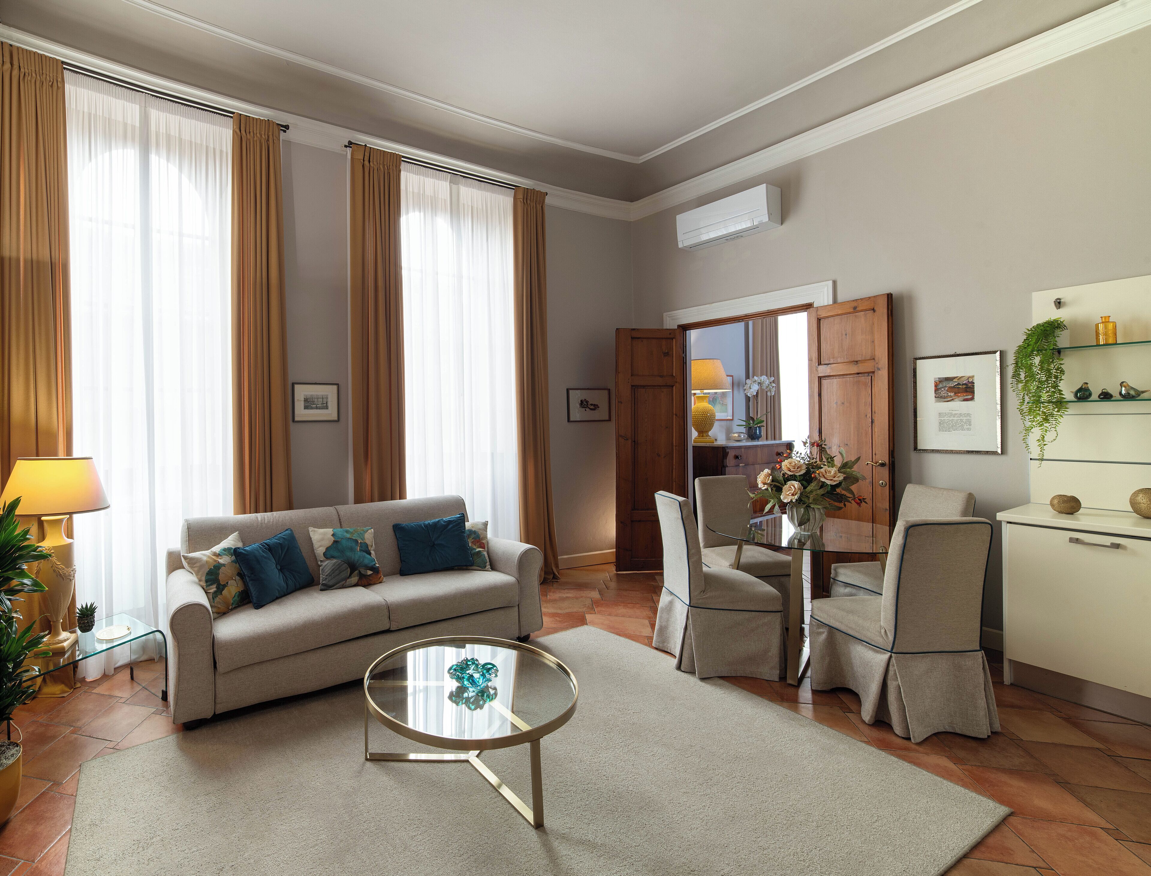 Ein Wohnzimmer mit einer Couch, einem Glastisch, einem Esstisch mit Stühlen sowie einer Küchenzeile inklusive Kühlschrank und Regalen.