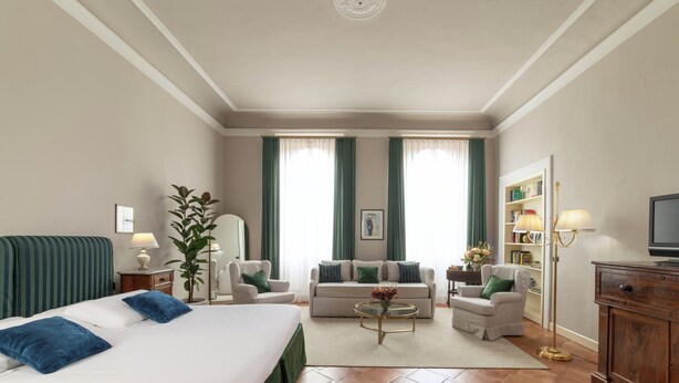 A hotel room with a bed, two armchairs, a sofa, a coffee table, a TV, a bookshelf, and a large window with curtains. - Palazzo Alfani al David (Florence)