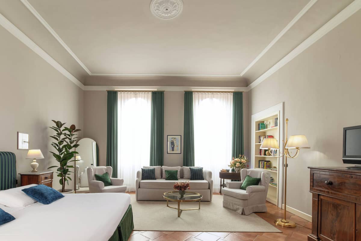 A hotel room with a bed, two armchairs, a sofa, a coffee table, a TV, a bookshelf, and a large window with curtains.