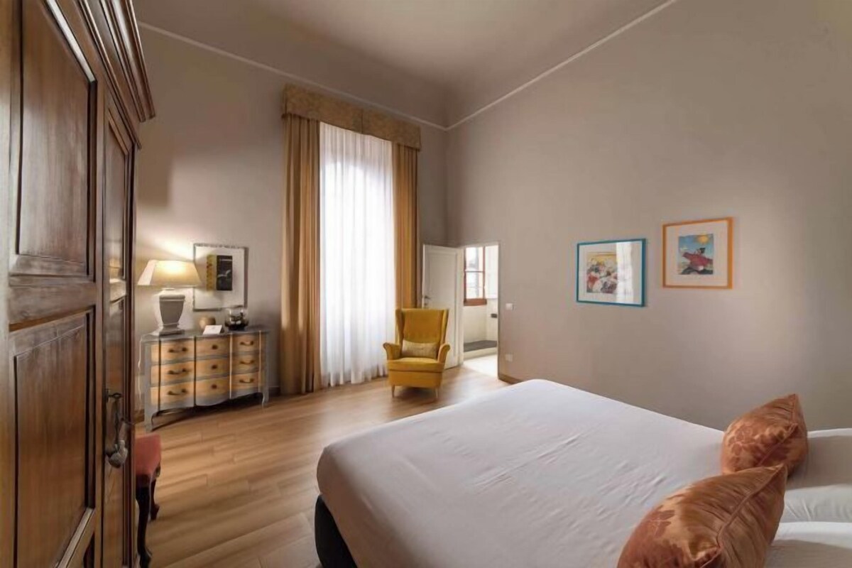 A hotel room with a large bed, a wooden wardrobe, a yellow armchair, a bedside table with a lamp, and two framed pictures on the wall.