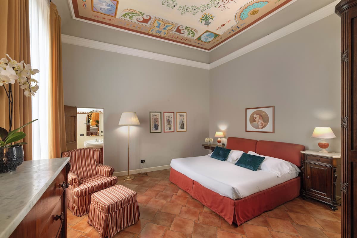 A hotel room with a large bed, striped armchair, and ottoman, a floor lamp, and a ceiling with decorative artwork.