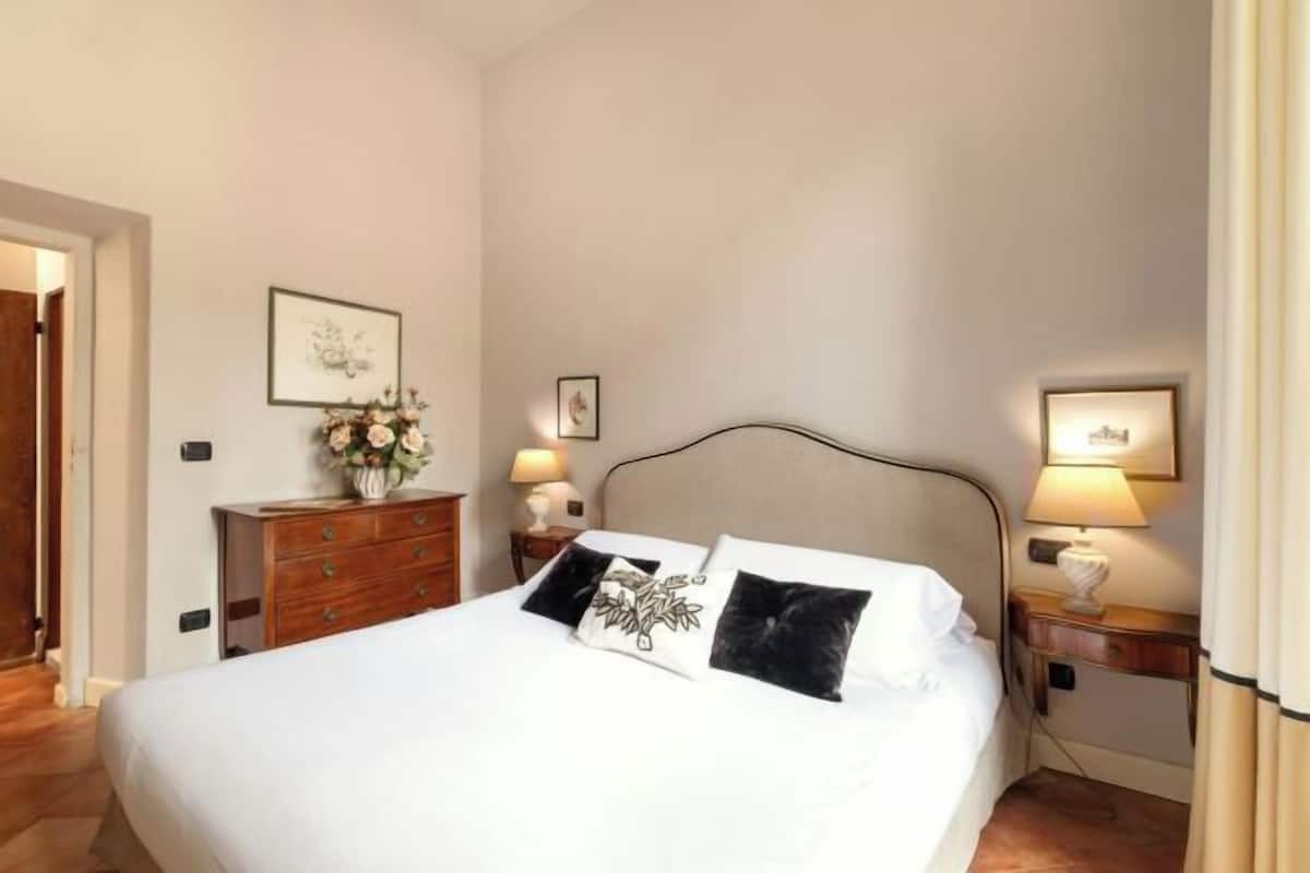 A neatly made bed with white linens and black and white patterned pillows, a wooden dresser with a vase of flowers, and two bedside lamps on wooden nightstands.