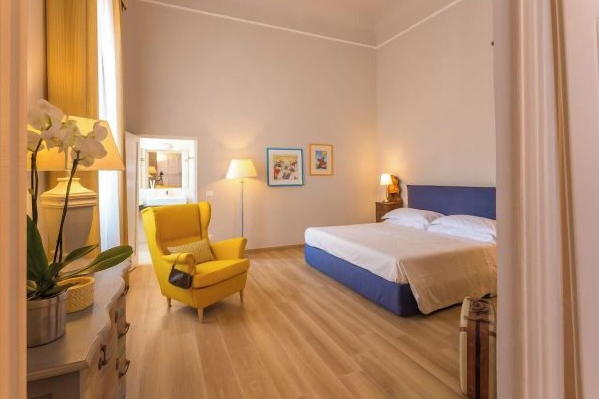 Une chambre d’hôtel avec un lit, un fauteuil jaune, une table de chevet, une lampe et un tableau au mur.