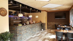 A bar area with a stone counter, shelves with bottles, and hanging lights. There are tables and chairs in the dining area with a patterned wall and a flat-screen TV mounted above. - Hotel im Rheintal (Kamp-Bornhofen)