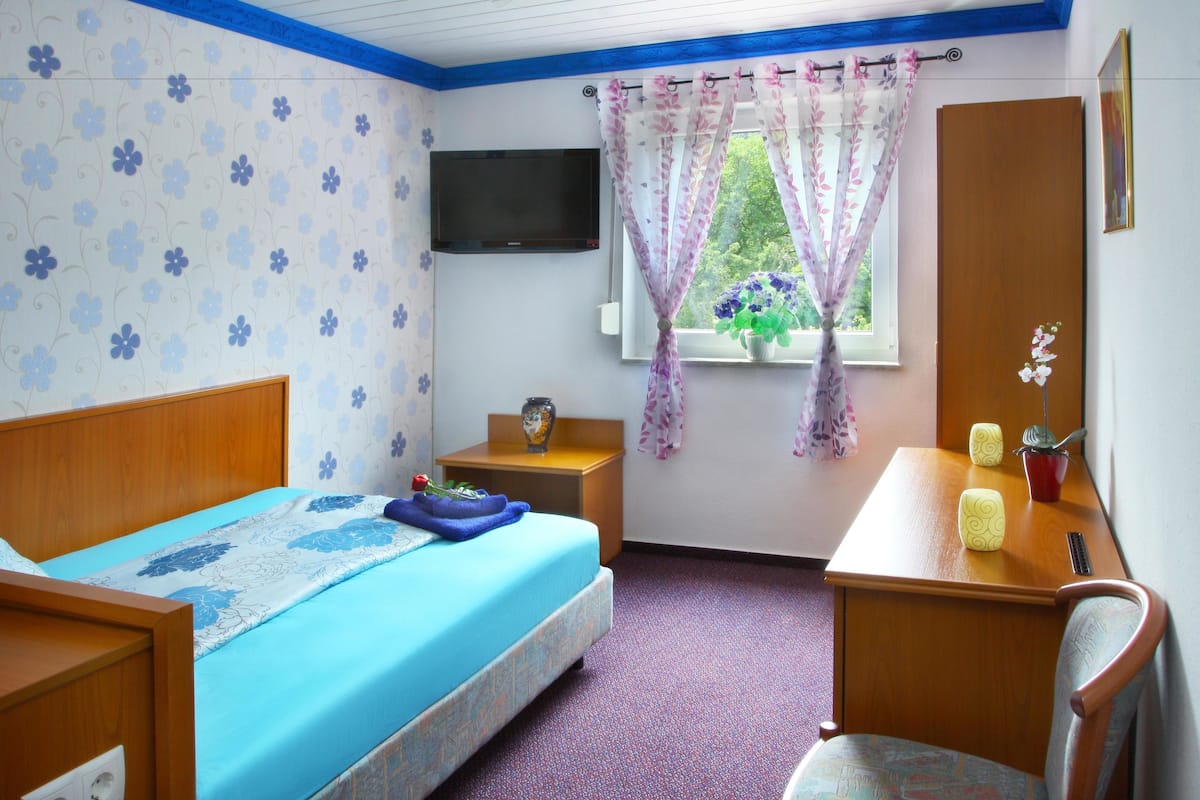 A bedroom with a bed, a desk, a chair, a TV, a window with curtains, and a wall-mounted TV.