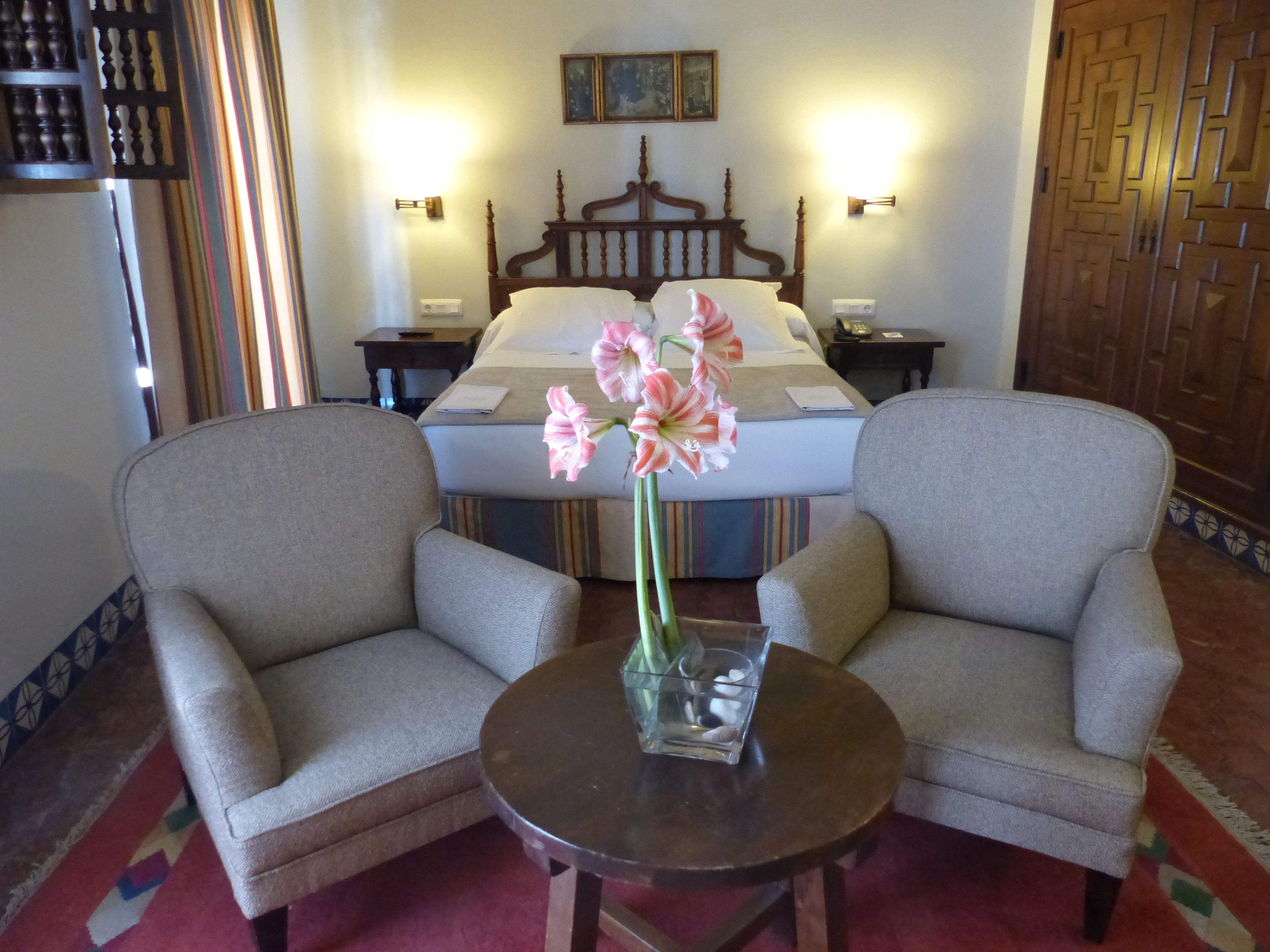 Una habitación de hotel con una cama, dos sillones, una mesita con un florero y una mesita de noche con una lámpara.