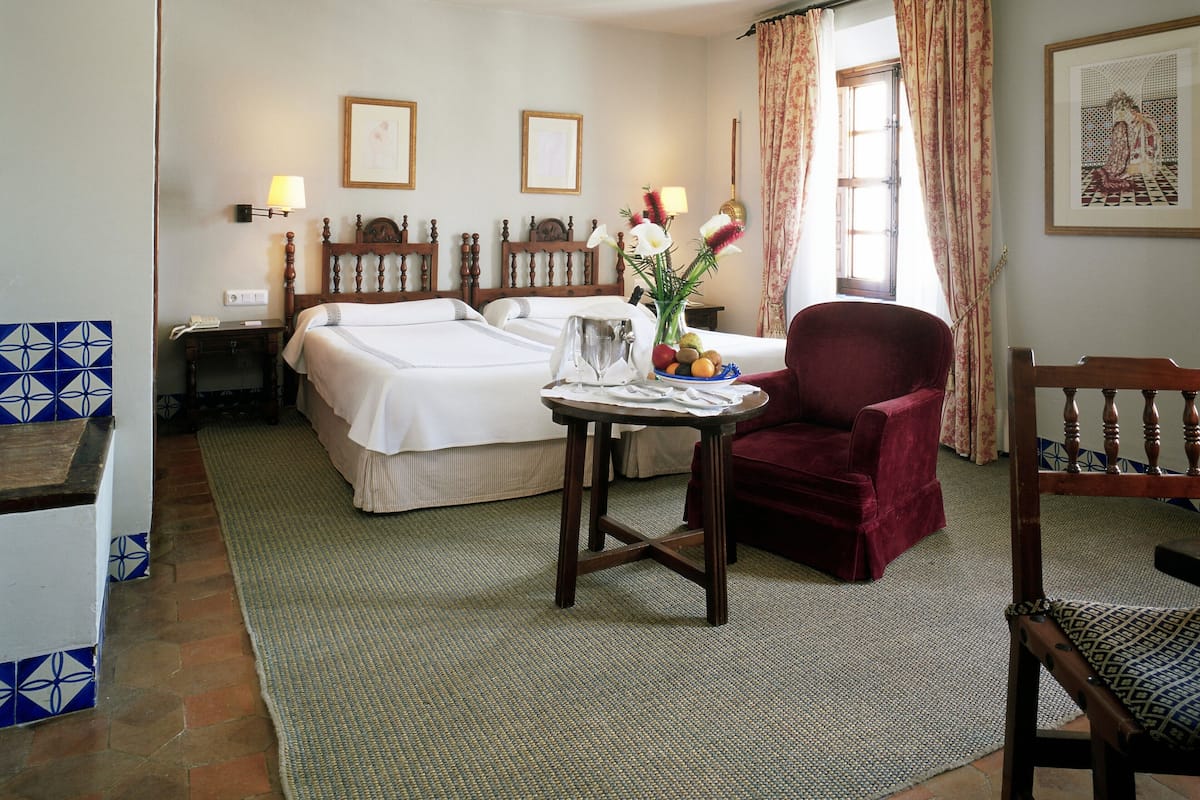 A hotel room with two beds, a dining table with a vase of flowers, a red armchair, and a framed picture on the wall.