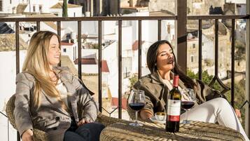Dos personas sentadas en sillas de mimbre en un balcón, con una botella de vino y copas sobre la mesa.