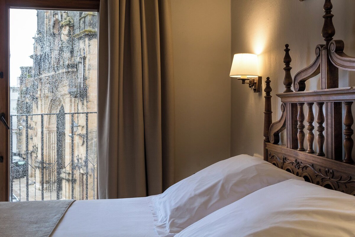 A bed with white linens, a wooden headboard, and a wall-mounted lamp. A window with curtains reveals a view of a building with statues.