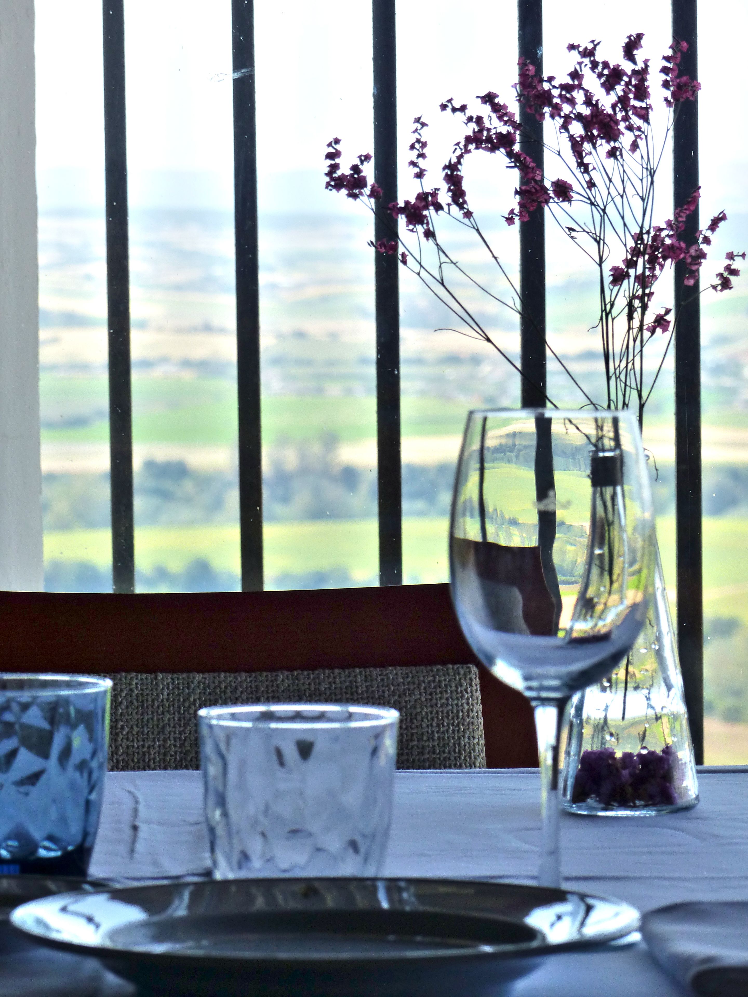 A table set with a clear glass vase containing purple flowers, a wine glass, and a patterned glass.