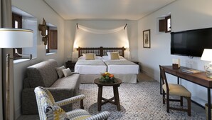 A hotel room with a double bed, a sofa, a small table with a flower arrangement, a desk with a chair, and a television.