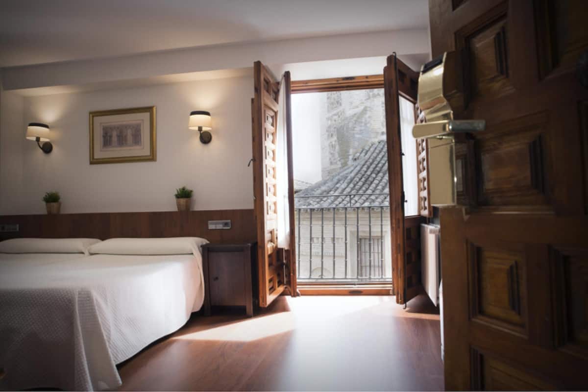 A hotel room with a bed, bedside tables, wall-mounted lamps, a window with a view of a building, and a wooden door.