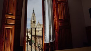 Groc a través d'una finestra amb estructura de fusta i cortina blanca, que mostra un edifici històric amb una torre campanar.