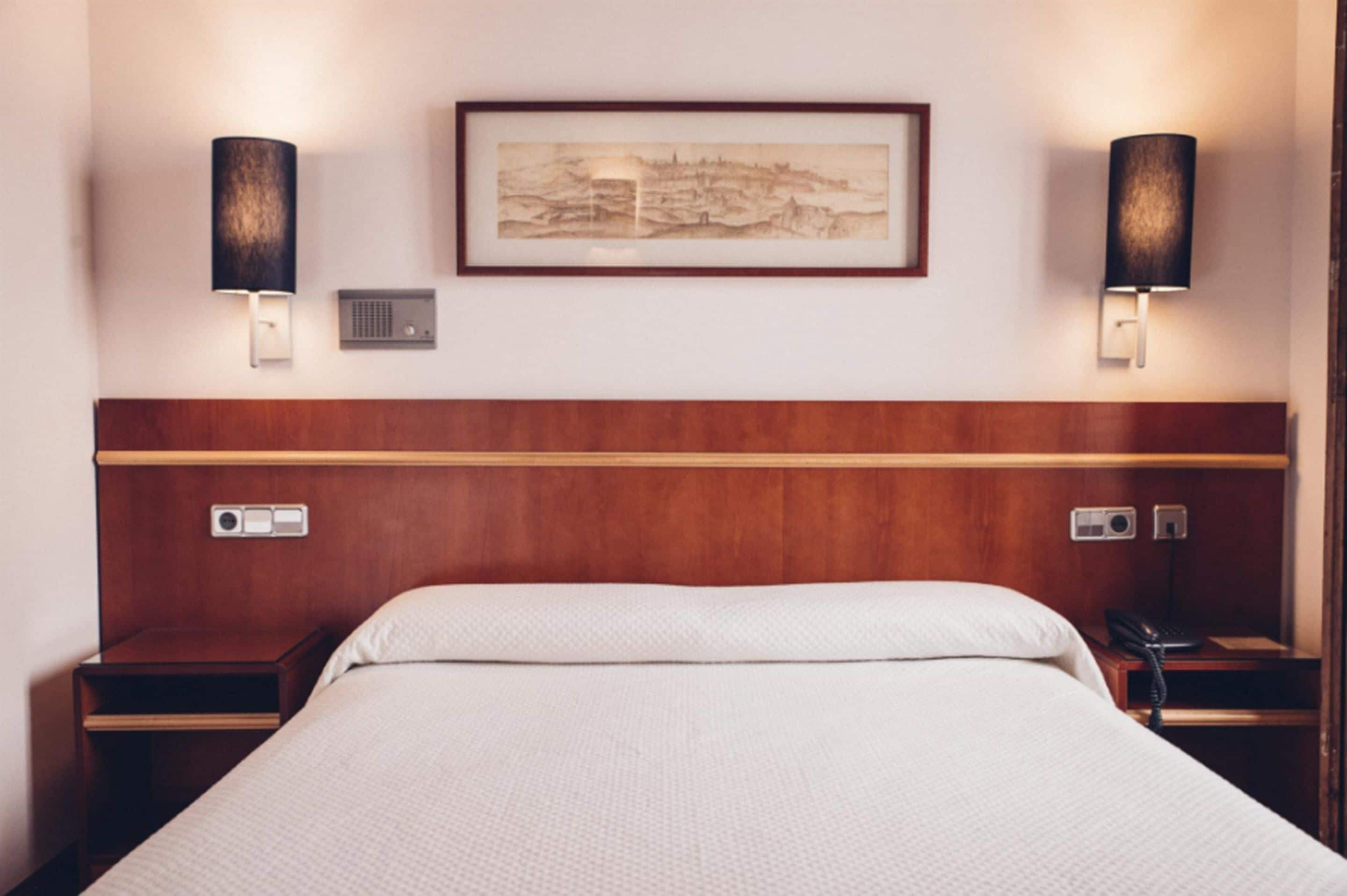 A hotel room with a wooden headboard, a bed with white bedding, two bedside tables with lamps, a framed artwork on the wall, and a telephone on one of the tables.