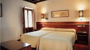 A hotel room with two beds, a wooden headboard, bedside lamps, a painting on the wall, and a desk with a chair.