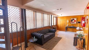 A modern hotel lobby with a black sofa, a small round table with a bottle, a decorative vase, and a fire extinguisher on the wall.