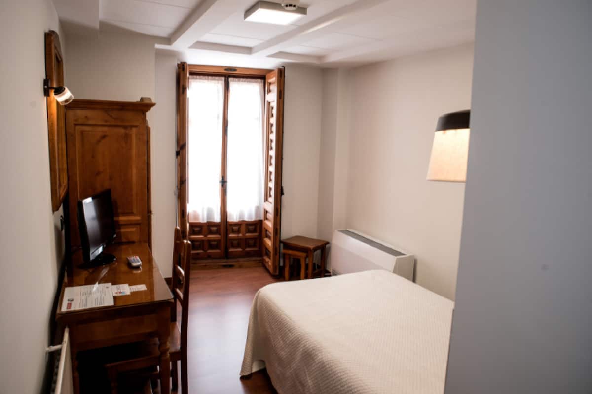 A hotel room with a bed, a desk with a computer, a chair, a TV, and a window with curtains.