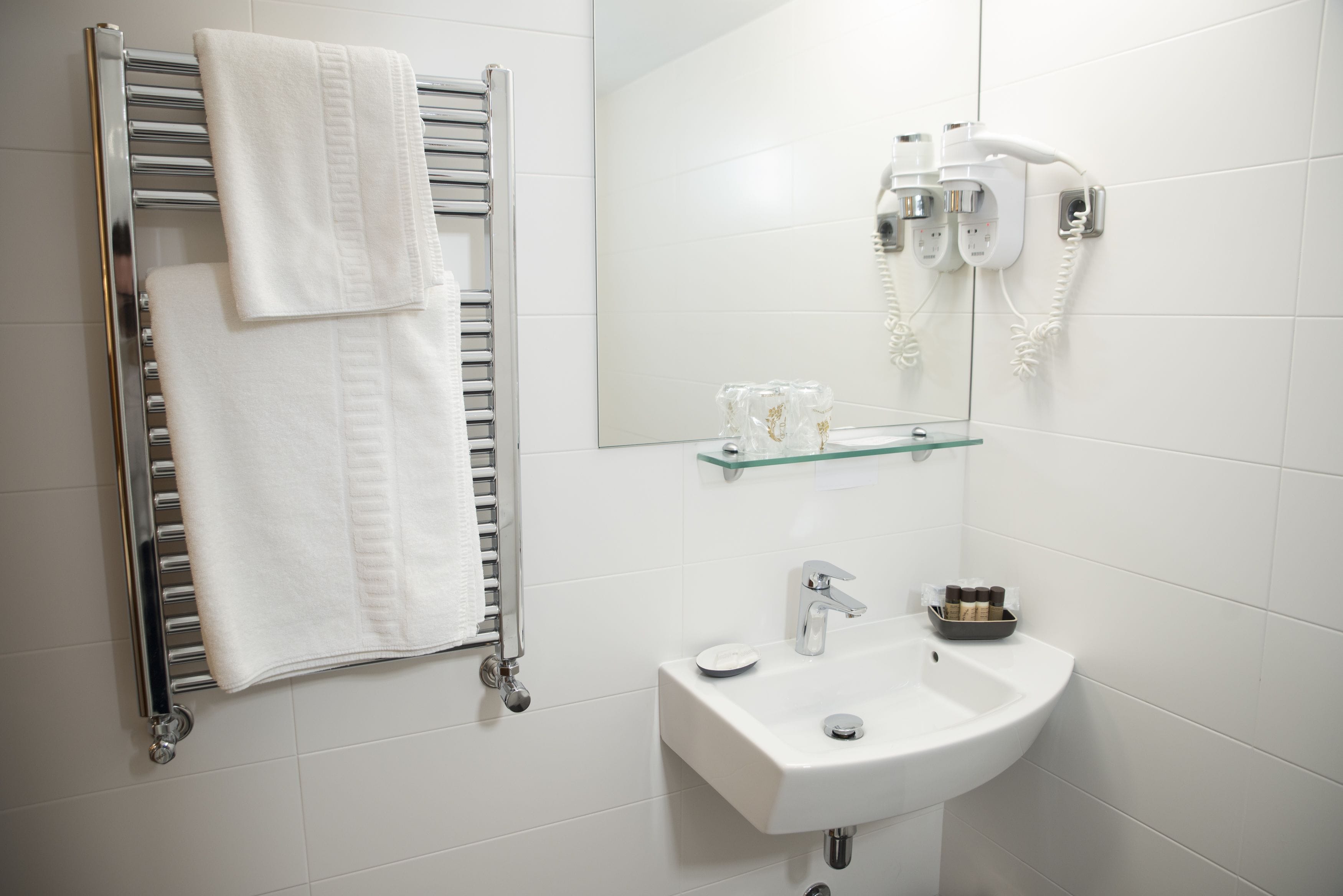 Un baño moderno con toallero cromado, repisa de vidrio con artículos de aseo, lavamanos blanco y secador de pelo montado en la pared.