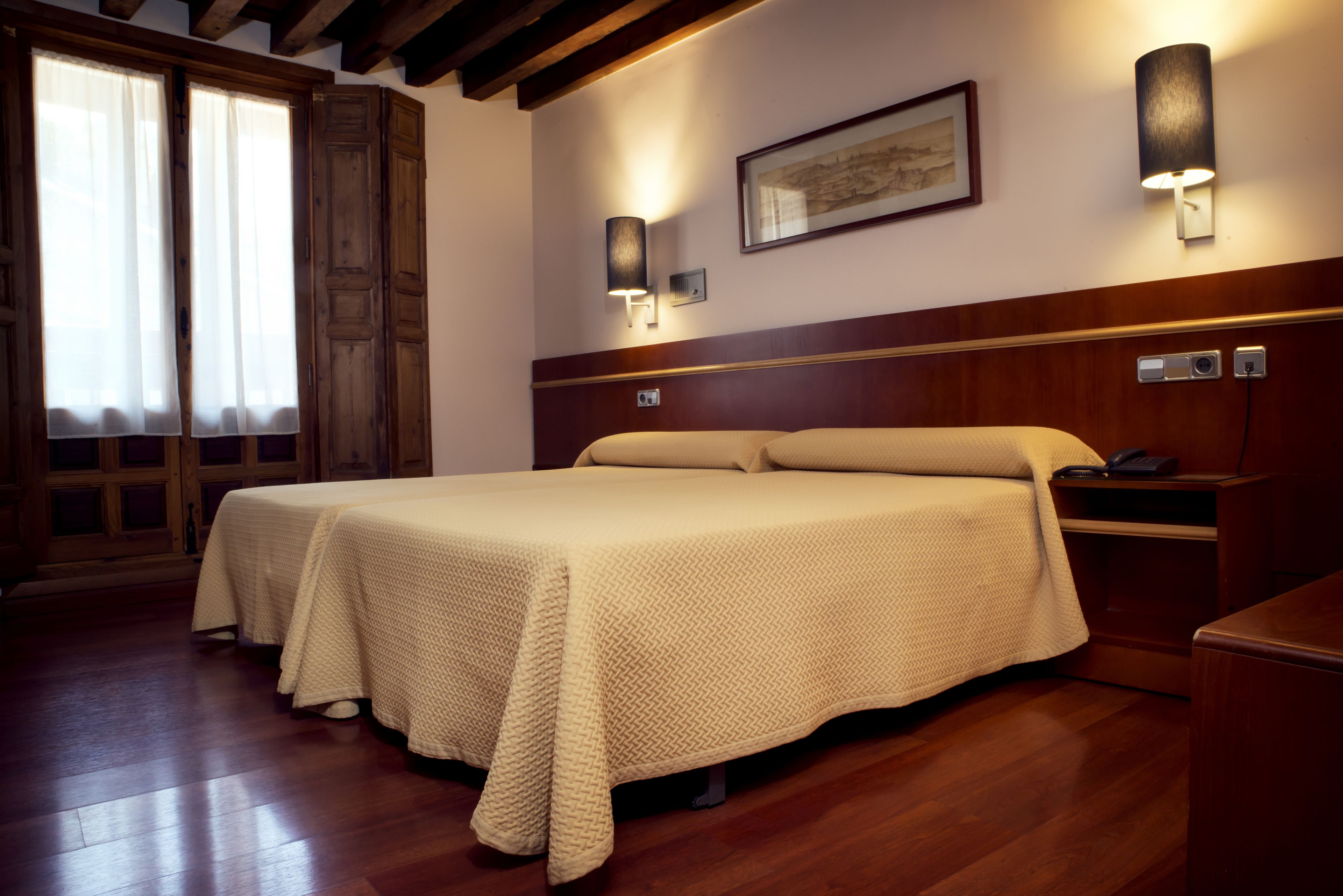 Una habitación de hotel con una cama grande, cabecera de madera, dos mesitas de noche, una mesita auxiliar y una lámpara fijada a la pared.