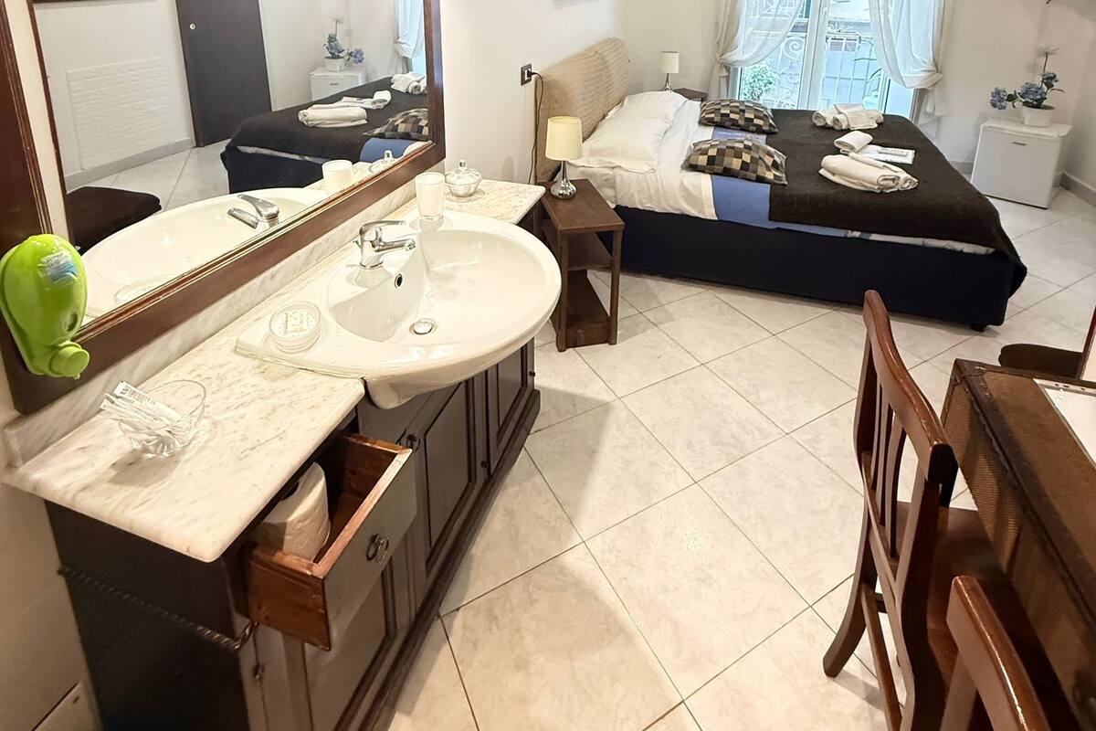 A hotel room with a large bed, a separate sitting area, a bathroom with a sink and mirror, and a window with curtains.