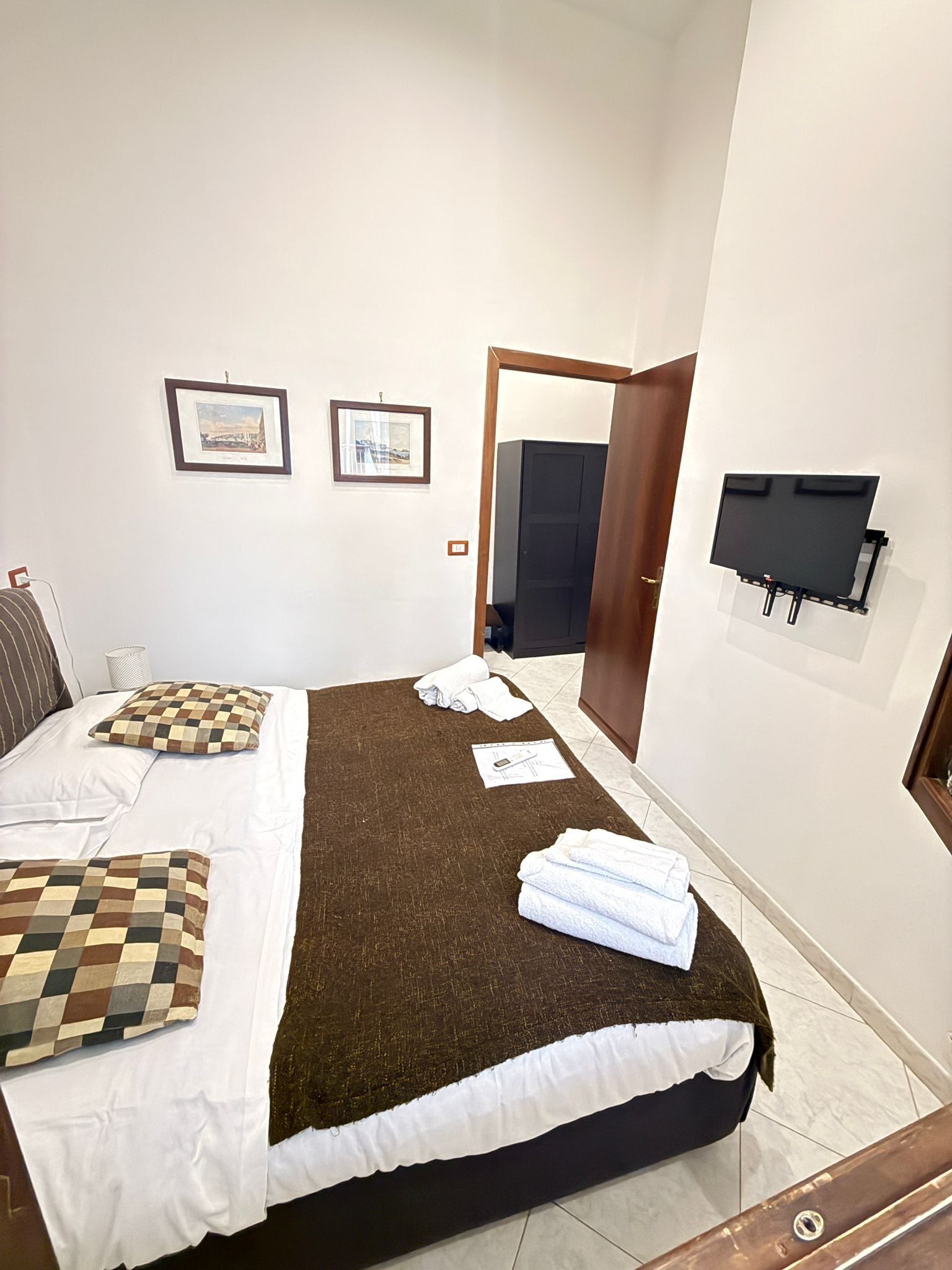 A neatly made bed with a checkered pillow, a brown blanket, and folded towels on a white mattress. There are two framed pictures on the wall, a door leading to another room, and a flat-screen TV mounted on the wall.