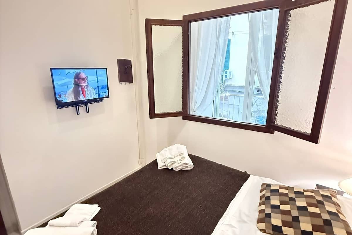 A hotel room with a bed, a TV, a window with curtains, and a view of buildings outside.