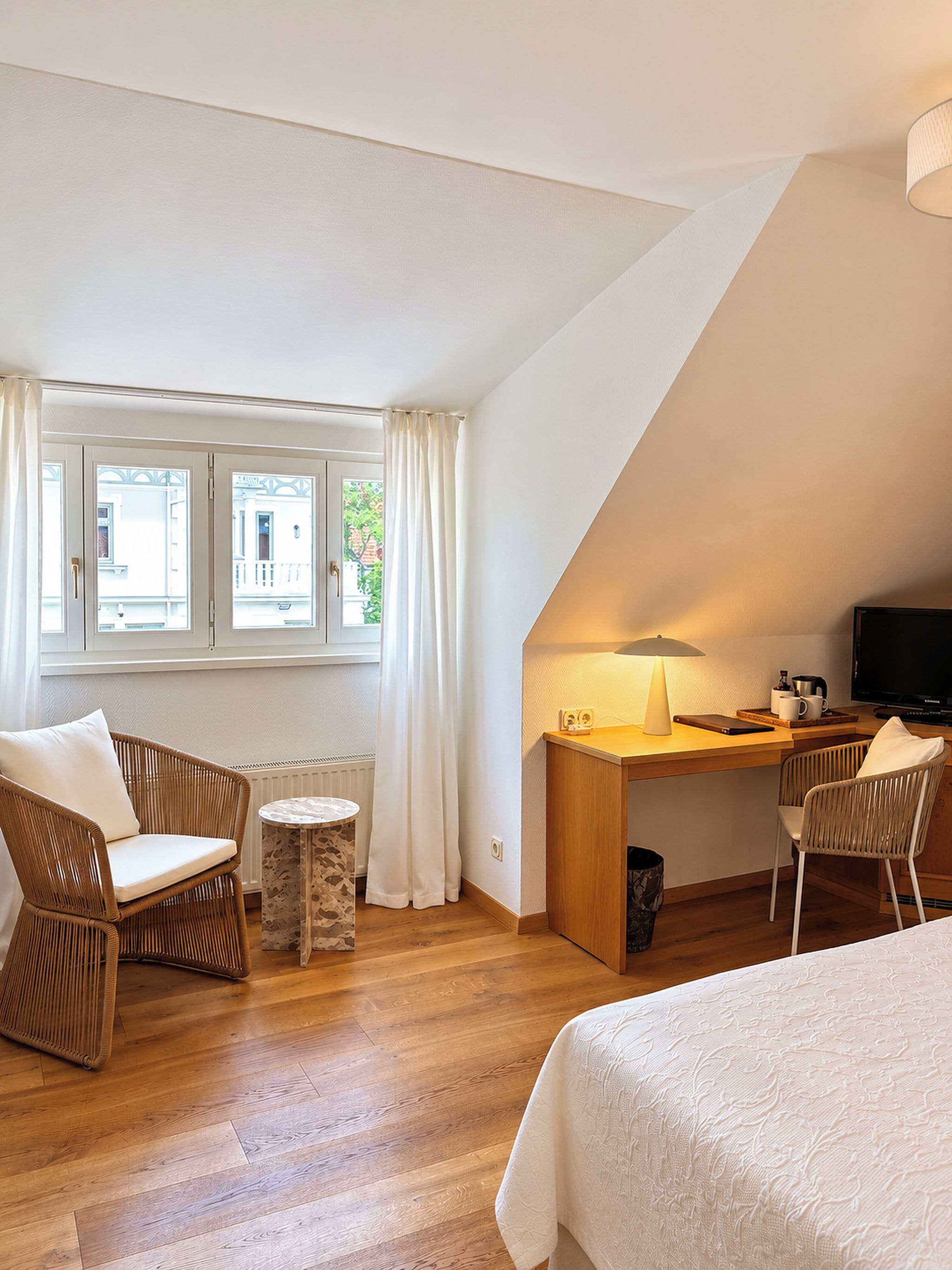 Ein modernes Hotelzimmer mit Holzboden, einem Schreibtisch mit Lampe, zwei Korbstühlen, einem Bett und Blick ins Freie durch ein Fenster mit Vorhängen.