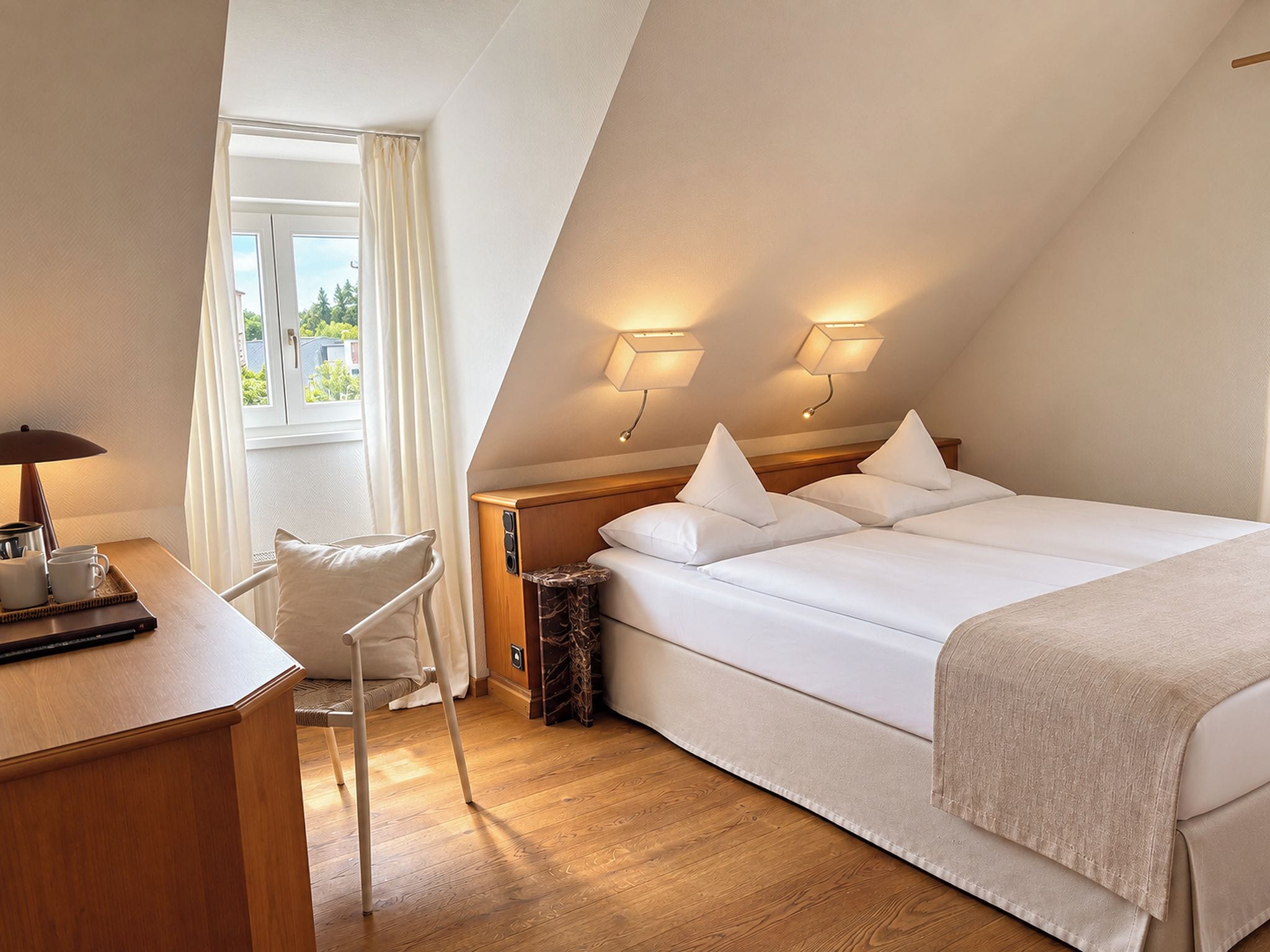 A hotel room with a bed, bedside tables, a desk with a chair, and a window with curtains.