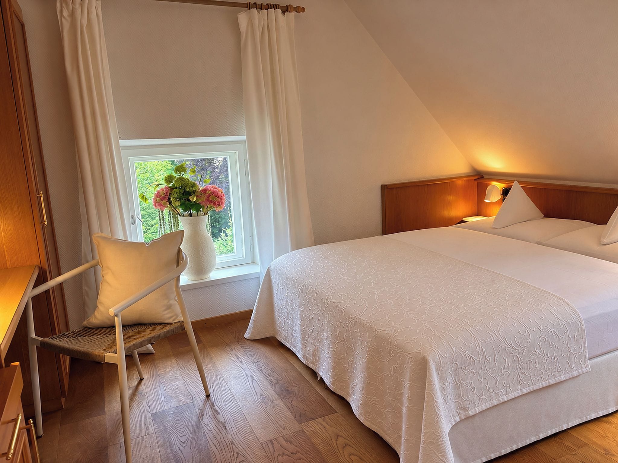 Een hotelkamer met een bed, een stoel, een raam met gordijnen en een vaas met bloemen.