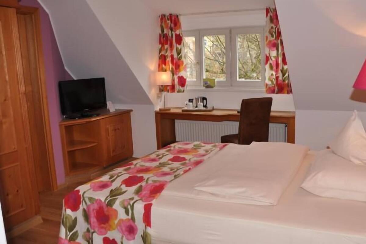 A bedroom with a bed, a desk, a chair, a television, and a window with curtains.