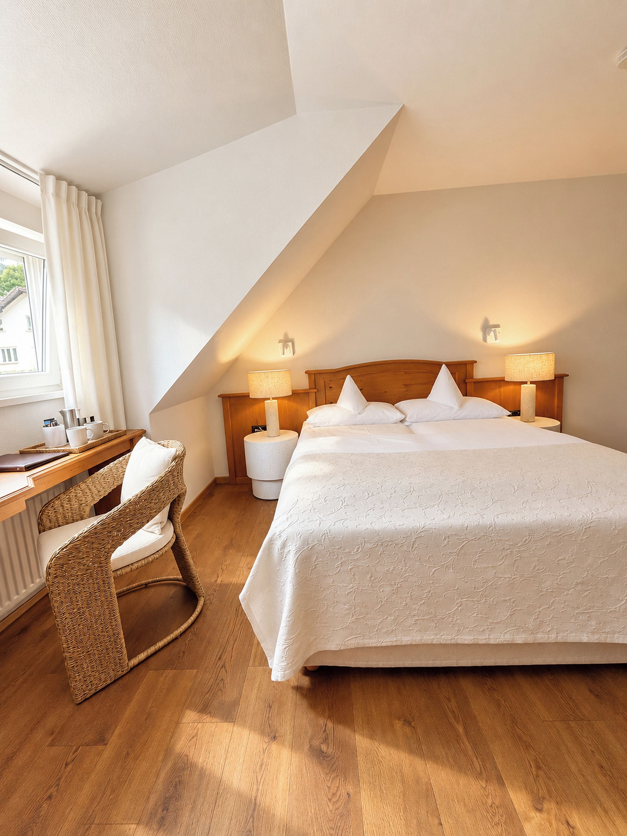 Een slaapkamer met een schuine kap, een bed met witte beddengoed, twee nachtlampjes, een rieten stoel, een houten bureau en een raam met gordijnen.