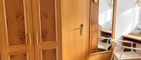 A room with a wooden wardrobe, a mirror, a chair, and a desk.