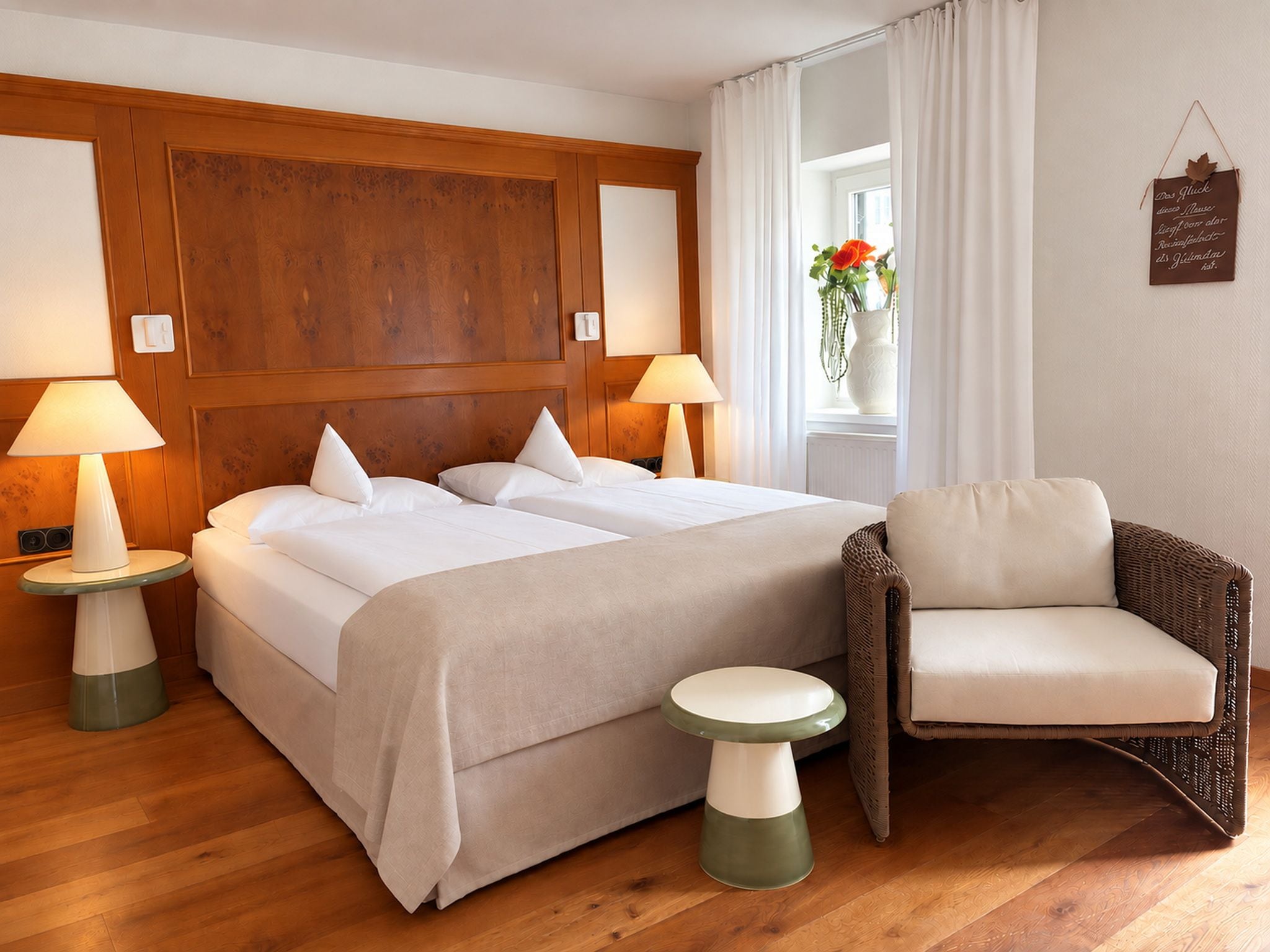 Une chambre d’hôtel avec une tête de lit en bois, deux lits avec du linge de lit blanc, deux lampes de chevet, un fauteuil en osier et une petite table.