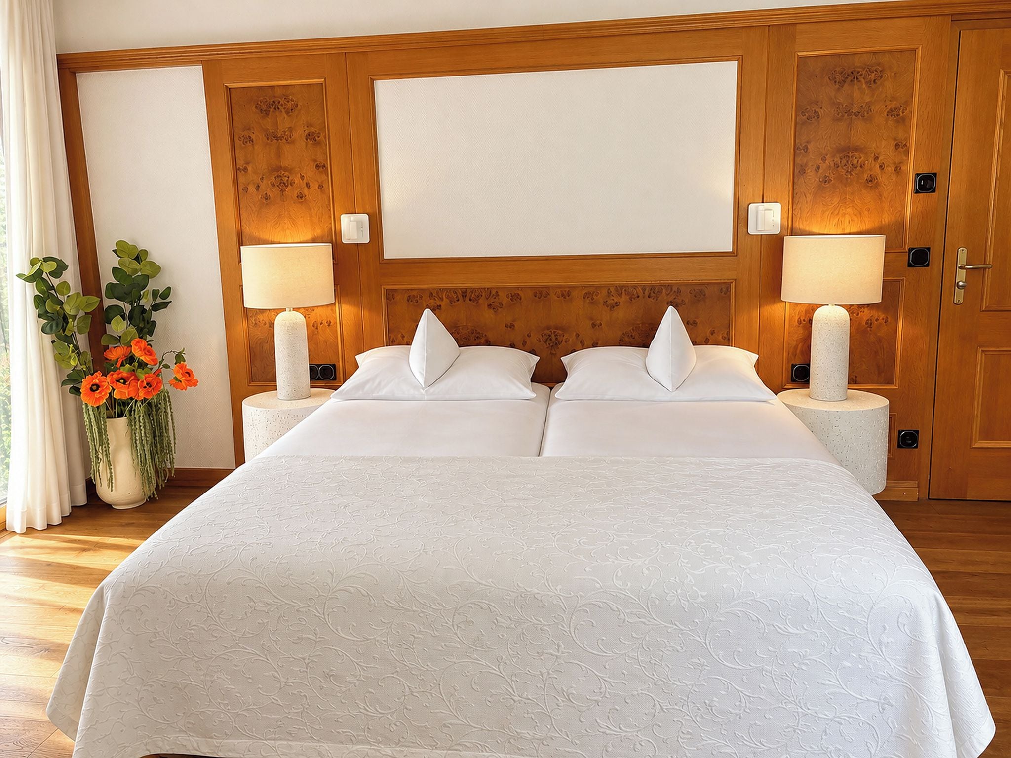 A double bed with a white bedspread, two bedside lamps, a wooden headboard, and a potted plant with orange flowers.