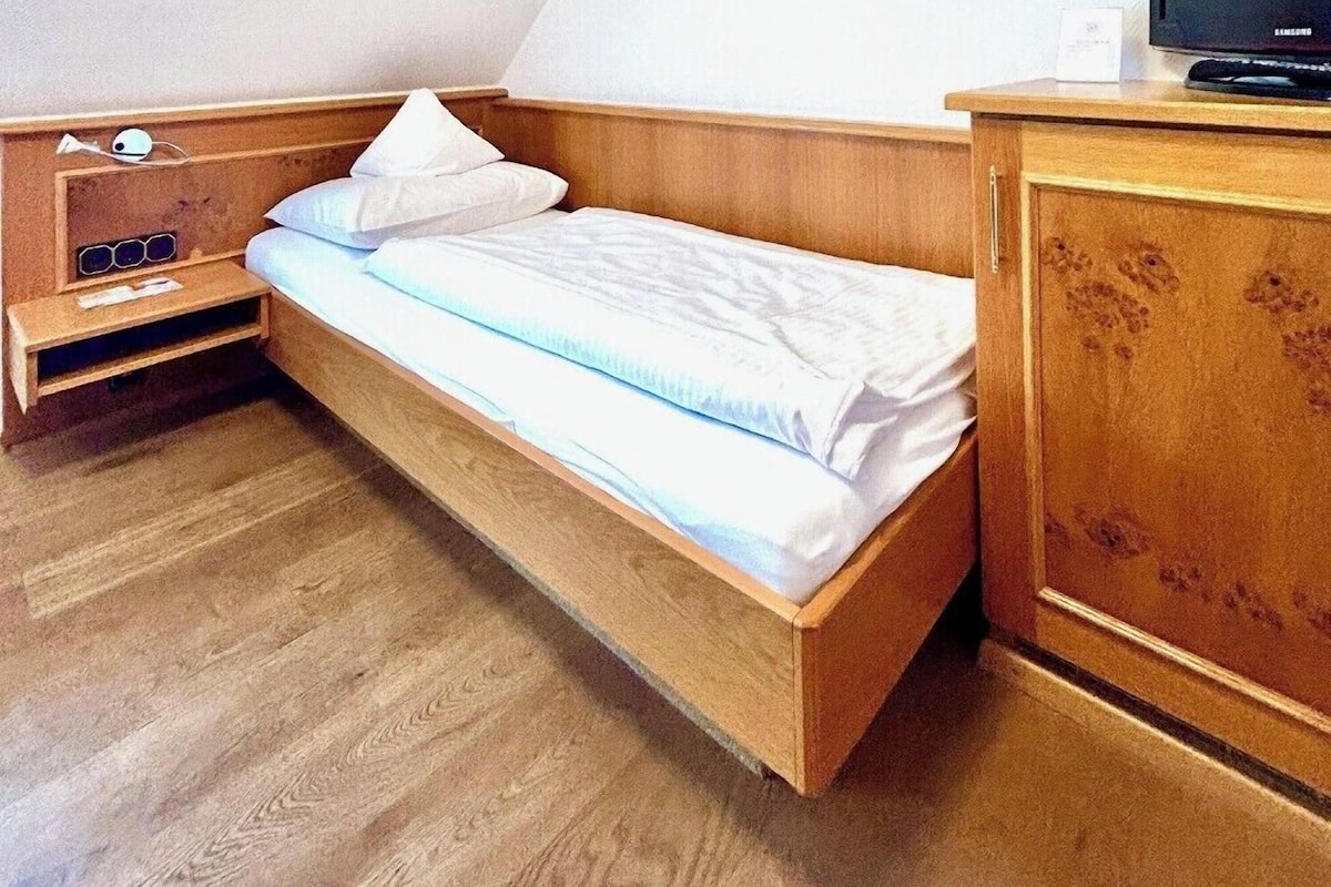 A small, single-bed room with a wooden headboard, a bedside table with a lamp, a television on a wooden cabinet, and a painting on the wall.
