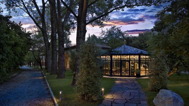 A gazebo with a glass enclosure, surrounded by a stone pathway and trees, set in a garden at dusk. - Ca' Mura' Hotel (Masera di Padova)