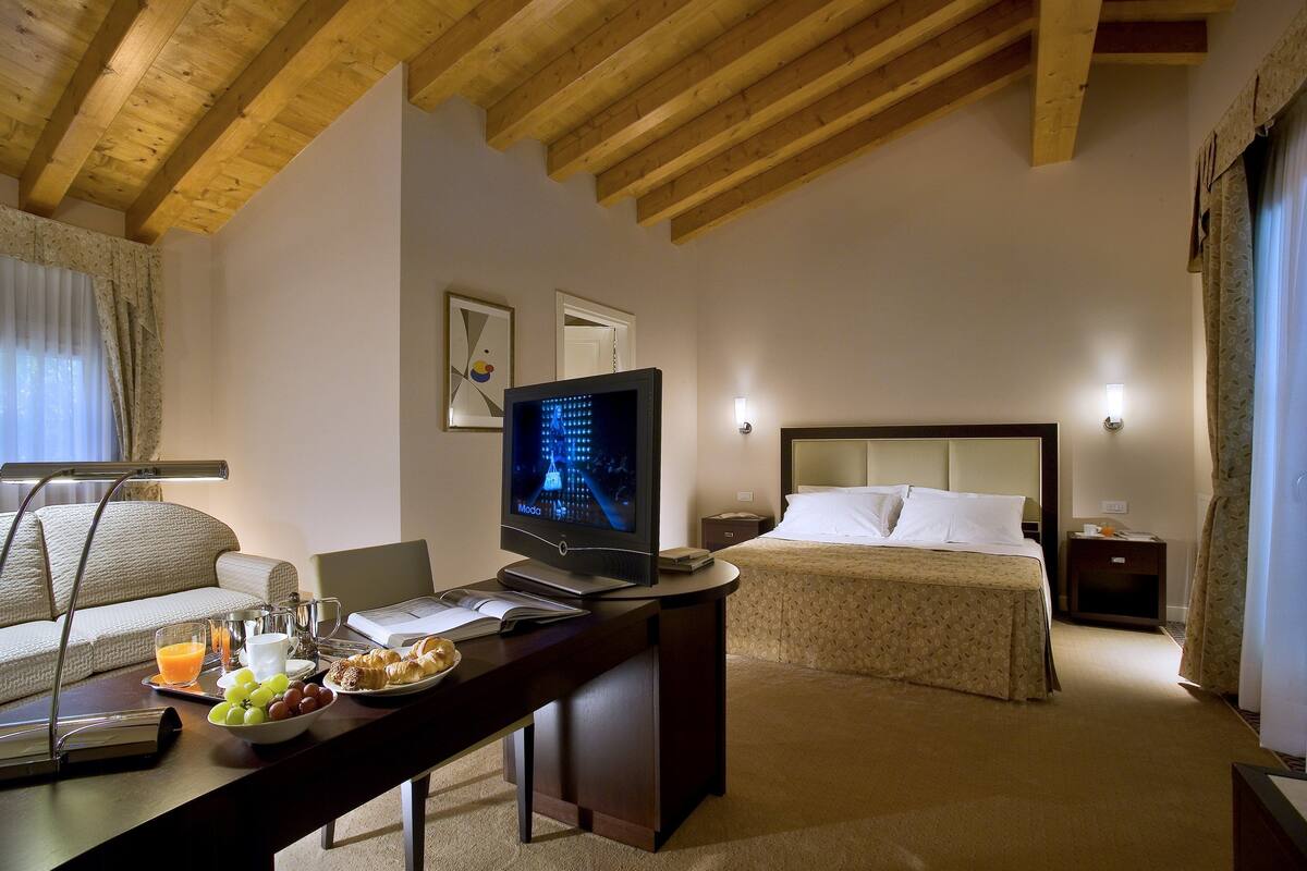 Habitación de hotel con techo de madera, una cama con cabecera, un escritorio con una laptop, un televisor y artículos de desayuno.